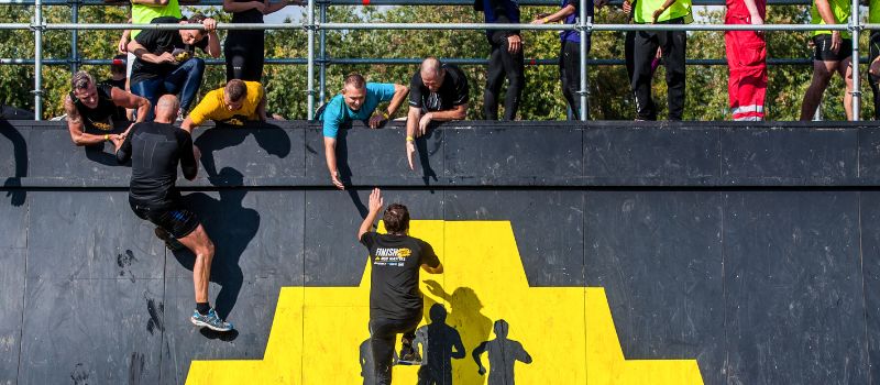 Deelnemers aan Mud Masters klimmen een steile wand op waarbij ze geholpen worden door anderen.